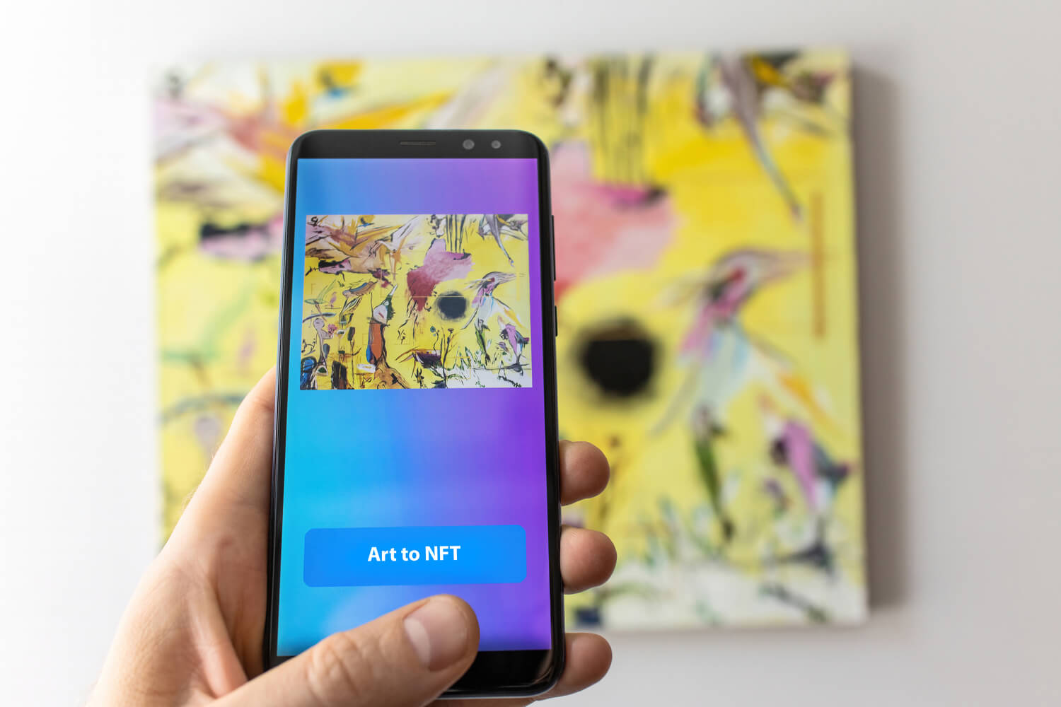 An image of a non-fungible token on a smartphone screen with a blue and purple background, and the physical piece of art behind the phone. 