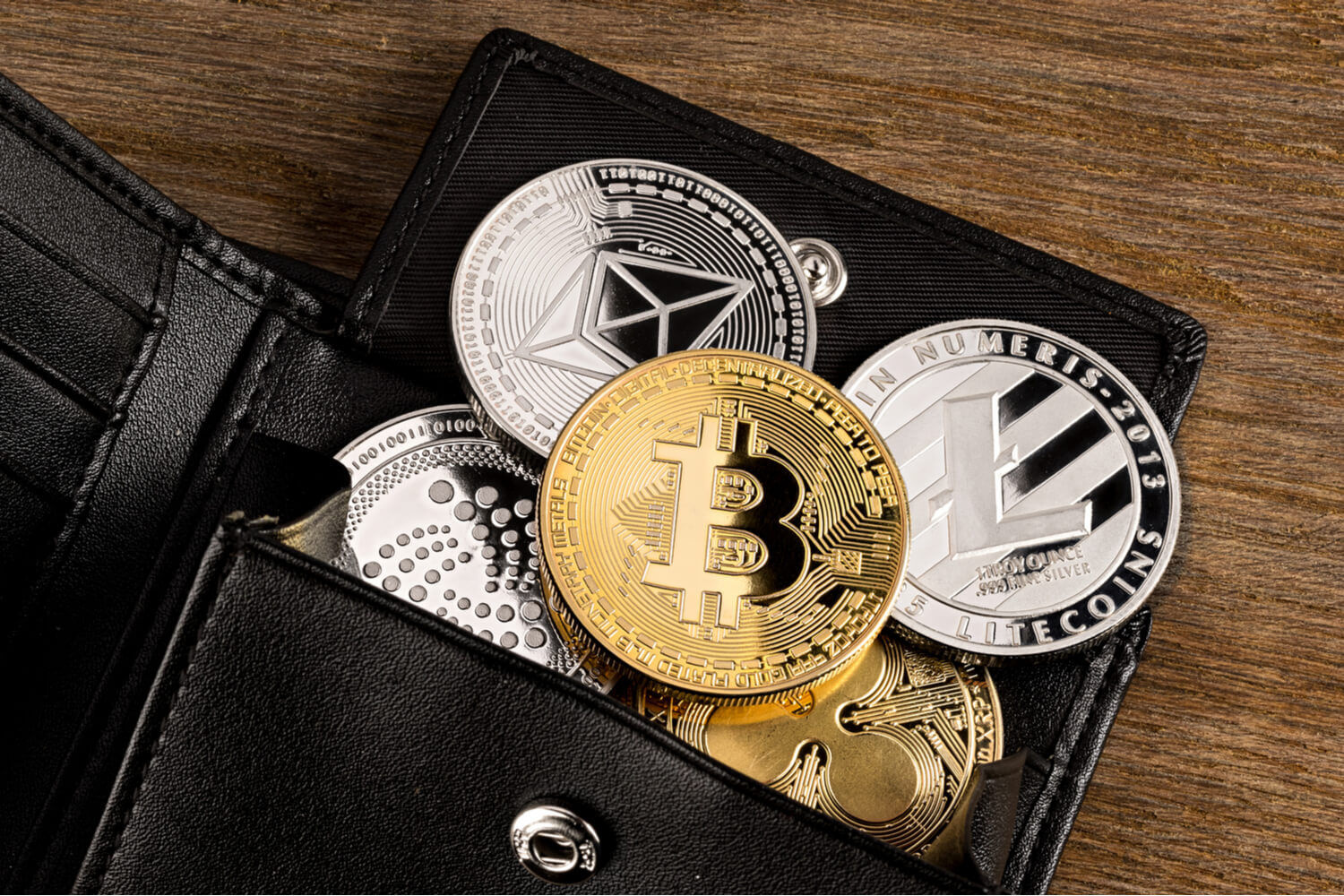A number of cryptocurrencies (such as Bitcoin, Ethereum and Litecoin), represented as physical coins, spilling out of a black leather wallet on a wooden background. 