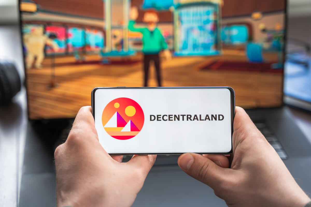 Logo of Decentraland shown on a mobile phone with the Decentraland metaverse projected onto a computer screen in the background.