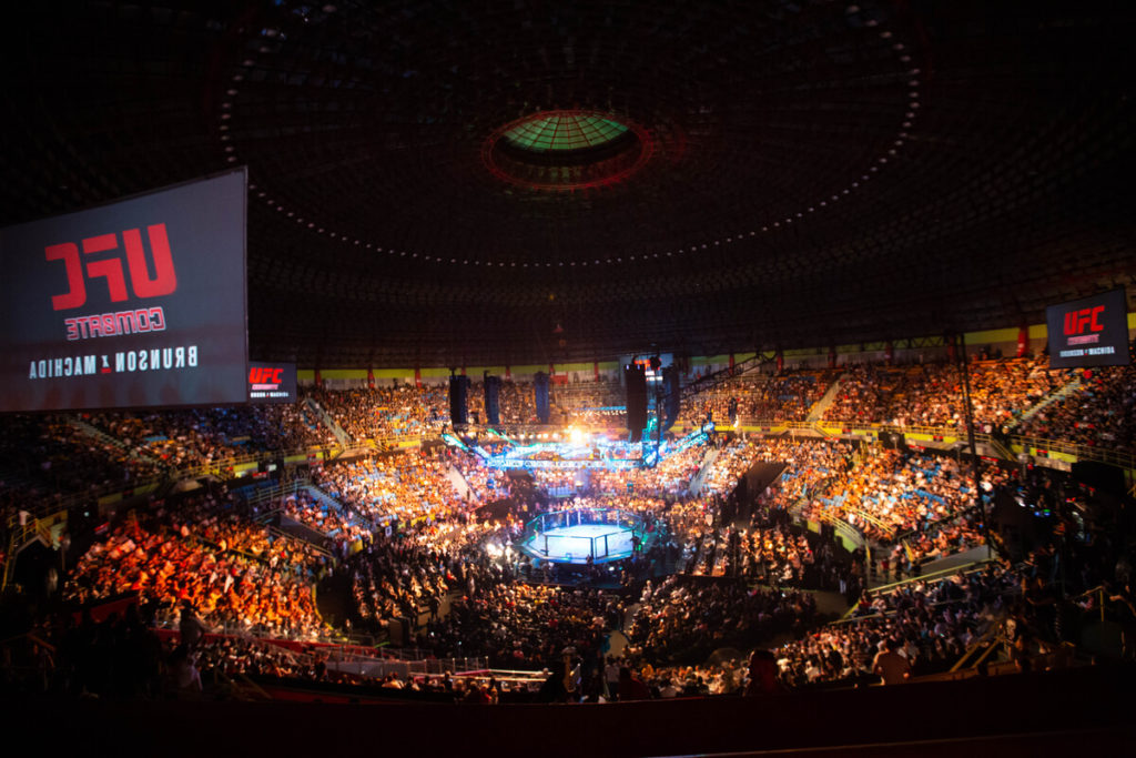 A crowded, big arena with an empty fighting ring in the middle