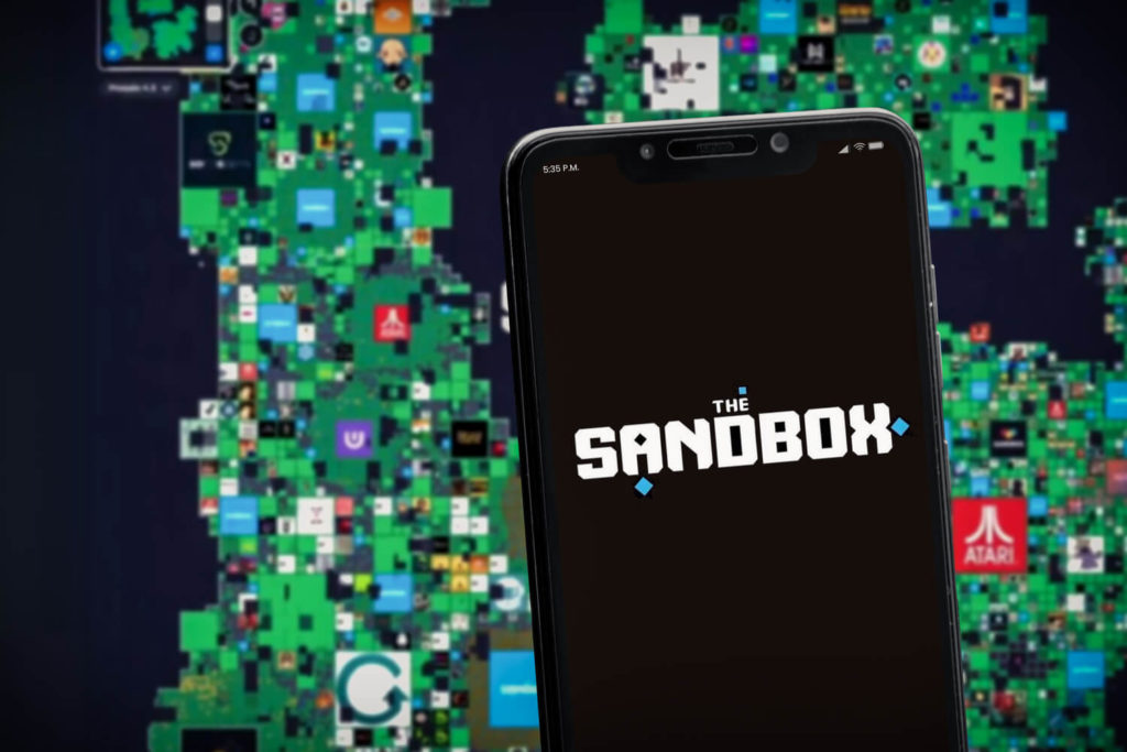 The Sandbox logo on a smartphone screen situated in front of an image of the Sandbox metaverse