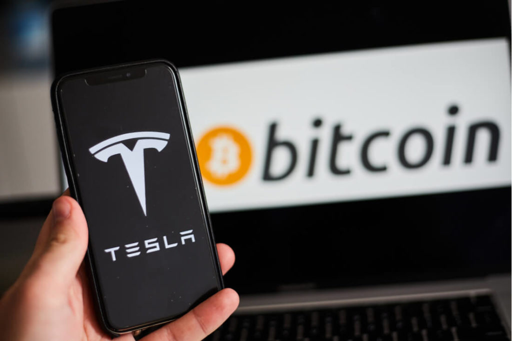 a mobile phone holding up Tesla logo with a laptop in the background showing Bitcoin logo