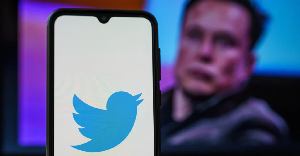 Image of the Twitter logo shown on an iPhone with Elon Musk sitting behind
