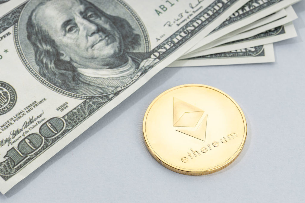 One Ethereum, represented in physical gold coin form, next to a pile of US dollars.