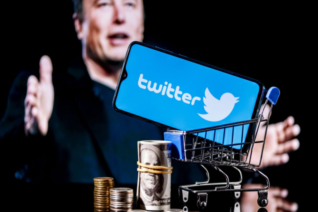 A small shopping cart with Twitter logo and US dollars and bills next to it with Elon Musk in the background