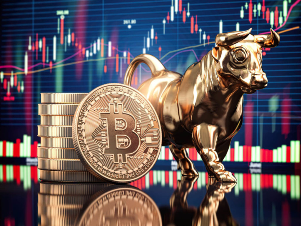 A bull figurine next to a pile of Bitcoins, represented in gold coin form, in front of a stock chart.