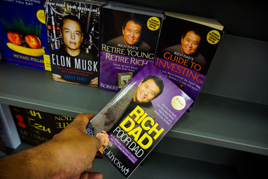 books by robert kiyosaki and elon musk on a shelf with rich dad poor dad being taken by a hand from the shelf