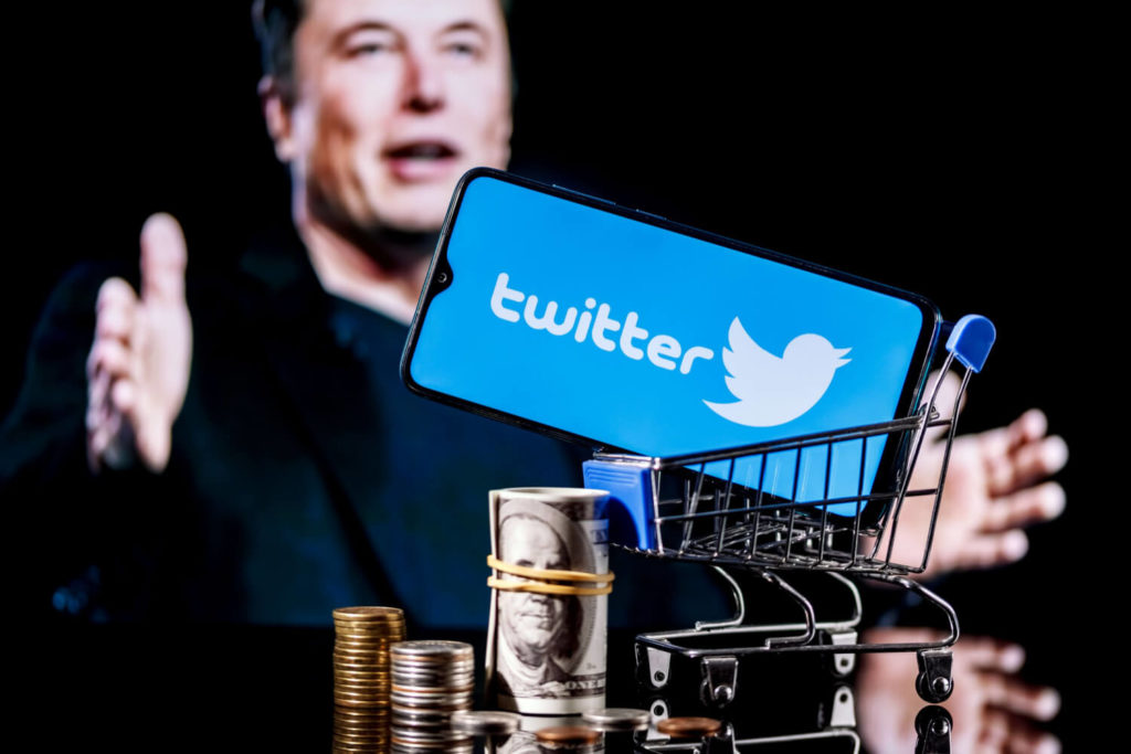Photograph of Elon Musk sitting behind a smartphone that is displaying the Twitter logo
