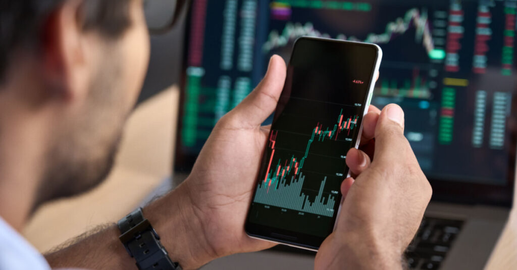 crypto trader trading on phone in front of pc screen with price charts